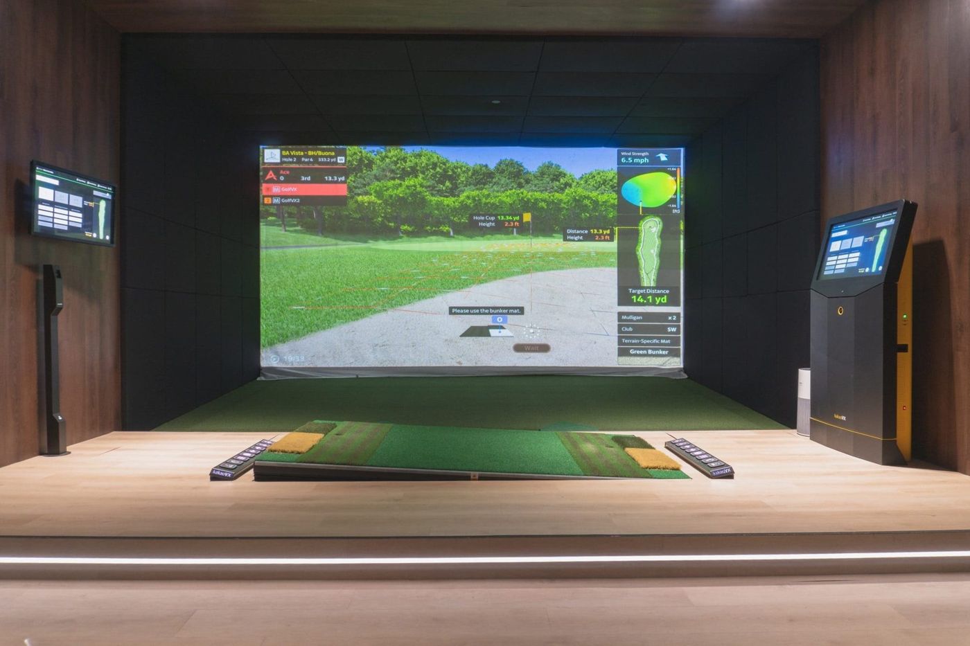 A Golf VX simulator bay with a wide screen, hitting mat and touchscreen kiosk in a warm wood-paneled room